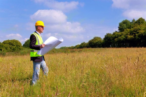 A man in yellow hard hat holding papers while standing on grass.
