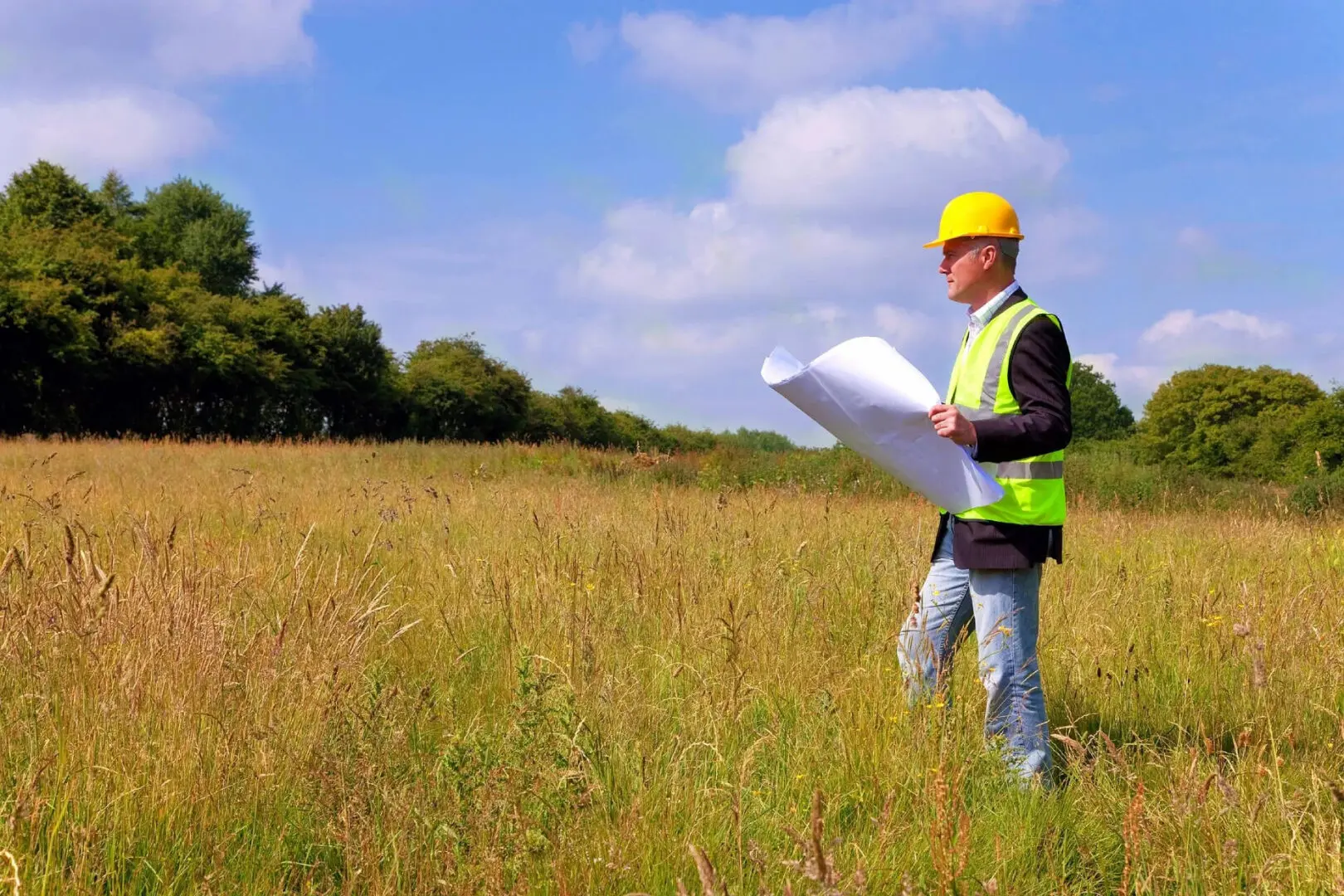 A man in yellow hard hat holding papers while standing on grass.